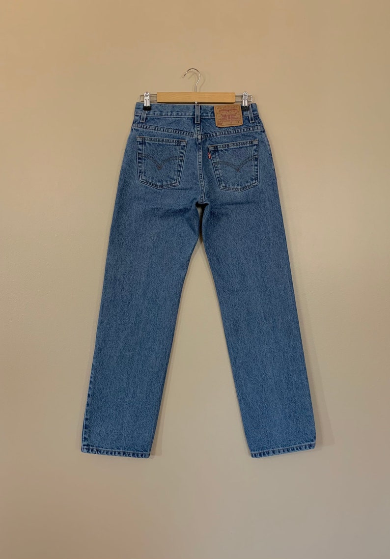 May include: Light blue denim jeans hanging on a wooden hanger. The jeans have a classic five-pocket design, a straight leg, and a brown leather patch on the back. The Levi's brand label is visible. The jeans are a medium wash.