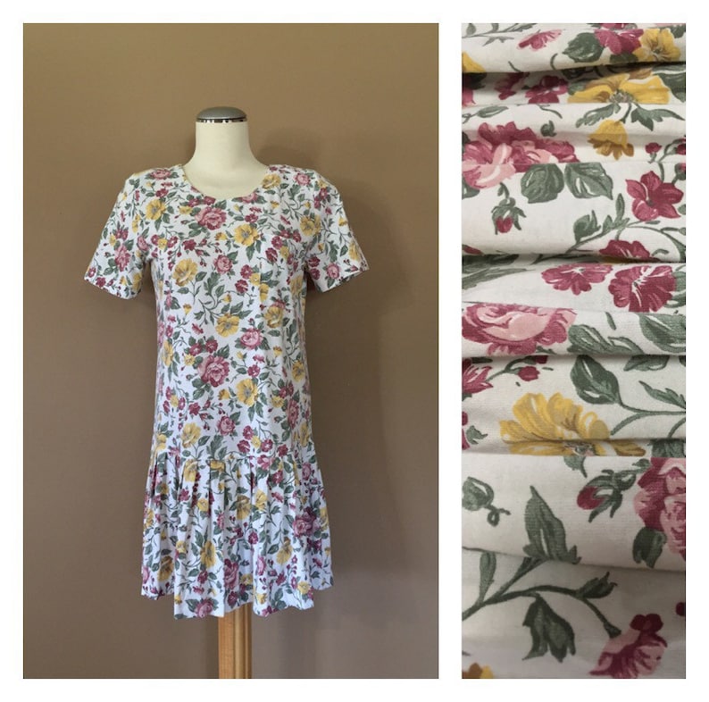 K&ouml;nnte beinhalten: Ein wei&szlig;es Kleid mit einem Blumendruck mit rosa, gelben und gr&uuml;nen Blumen. Das Kleid hat kurze &Auml;rmel und einen gefalteten Saum.