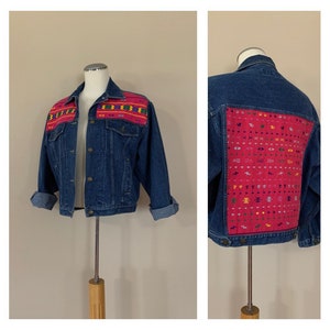 May include: A blue denim jacket with colorful embroidered designs on the back and shoulders. The embroidery features geometric patterns in pink, yellow, green, and blue.