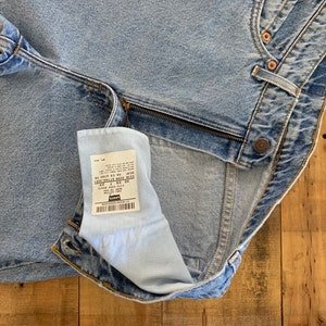 May include: Light blue denim jeans with a zipper and button closure. The jeans have a classic five-pocket design and a faded wash. A tag is visible inside the jeans. The jeans are laid out on a wooden surface.