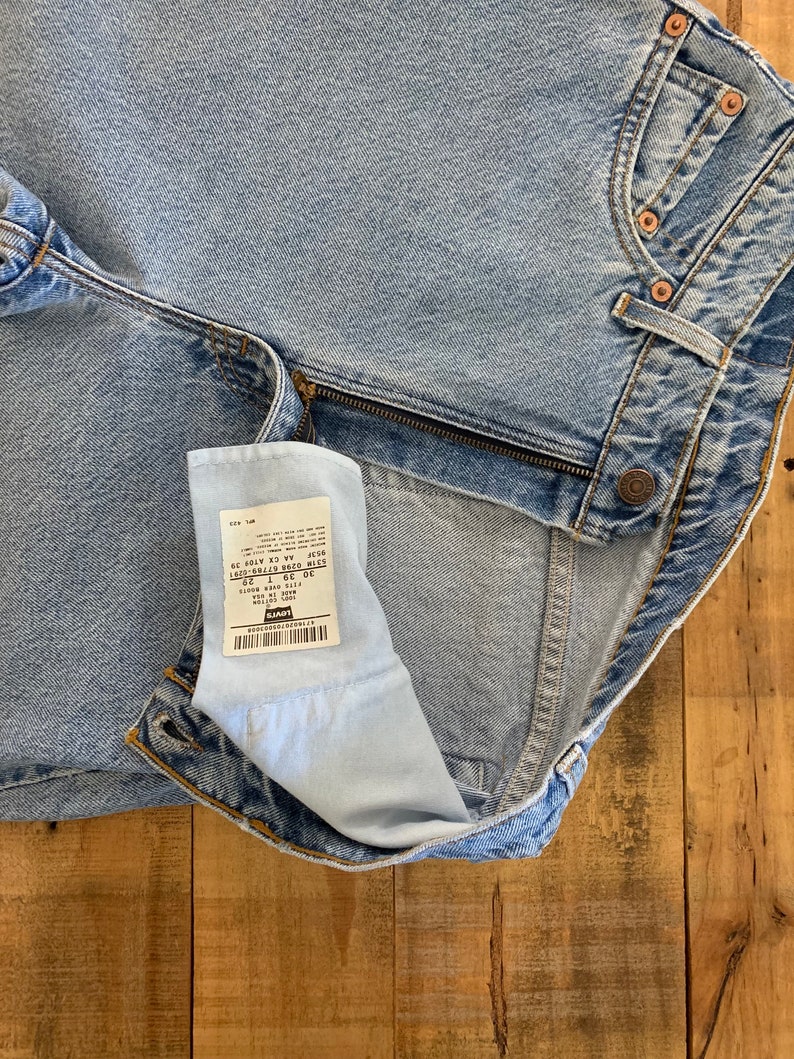May include: Light blue denim jeans with a zipper and button closure. The jeans have a classic five-pocket design and a faded wash. A tag is visible inside the jeans. The jeans are laid out on a wooden surface.