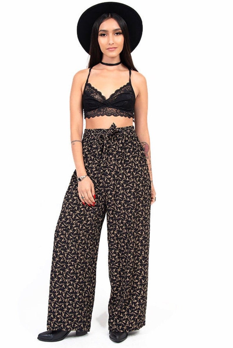 Puede incluir: Una mujer con un bralette de encaje negro y pantalones anchos con estampado floral negro y dorado. Tambi&eacute;n lleva un sombrero negro y zapatos negros.