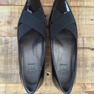 May include: A pair of black patent leather shoes with a black criss-cross strap across the vamp. The shoes are labeled "CIRCA Comfort" and "JOURNEY" on the inside.