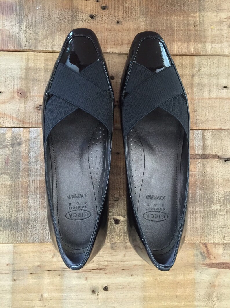May include: A pair of black patent leather shoes with a black criss-cross strap across the vamp. The shoes are labeled "CIRCA Comfort" and "JOURNEY" on the inside.