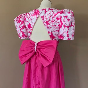 Puede incluir: Un vestido de estampado floral rosa y blanco con un gran lazo rosa en la espalda. El vestido tiene una abertura de ojo de cerradura en la espalda y mangas cortas abullonadas.