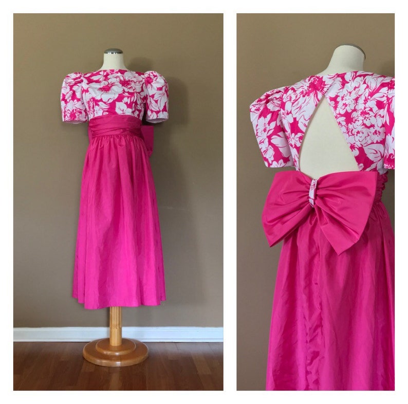 Puede incluir: Un vestido rosa con un corpi&ntilde;o de estampado floral blanco y un gran lazo rosa en la espalda. El vestido tiene mangas abullonadas y una falda completa.