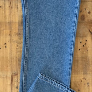 May include: Light blue denim jeans, folded at the hem, with visible stitching details. The jeans are laid flat on a wooden surface, showcasing the classic five-pocket design and a straight leg cut. The fabric has a medium wash.