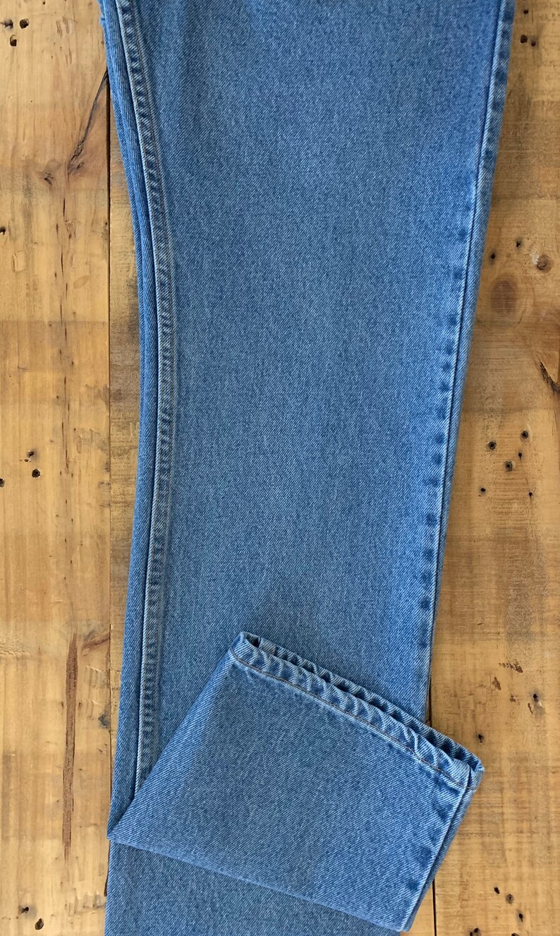 May include: Light blue denim jeans, folded at the hem, with visible stitching details. The jeans are laid flat on a wooden surface, showcasing the classic five-pocket design and a straight leg cut. The fabric has a medium wash.