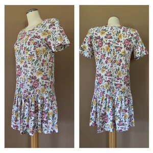 K&ouml;nnte beinhalten: Ein wei&szlig;es Kleid mit einem Blumendruck in Rosa-, Gelb- und Gr&uuml;nt&ouml;nen. Das Kleid hat kurze &Auml;rmel und einen gerafften Rock.
