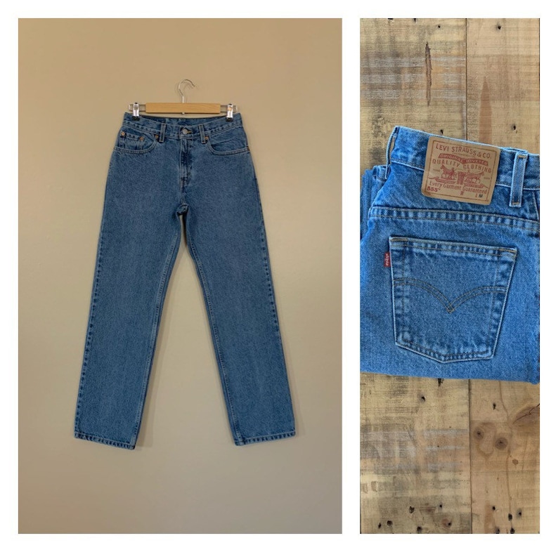 May include: Light blue denim jeans hanging on a wooden hanger. The jeans feature a classic five-pocket design and a button and zip closure. A close-up shows the back pocket with a red Levi's tag.