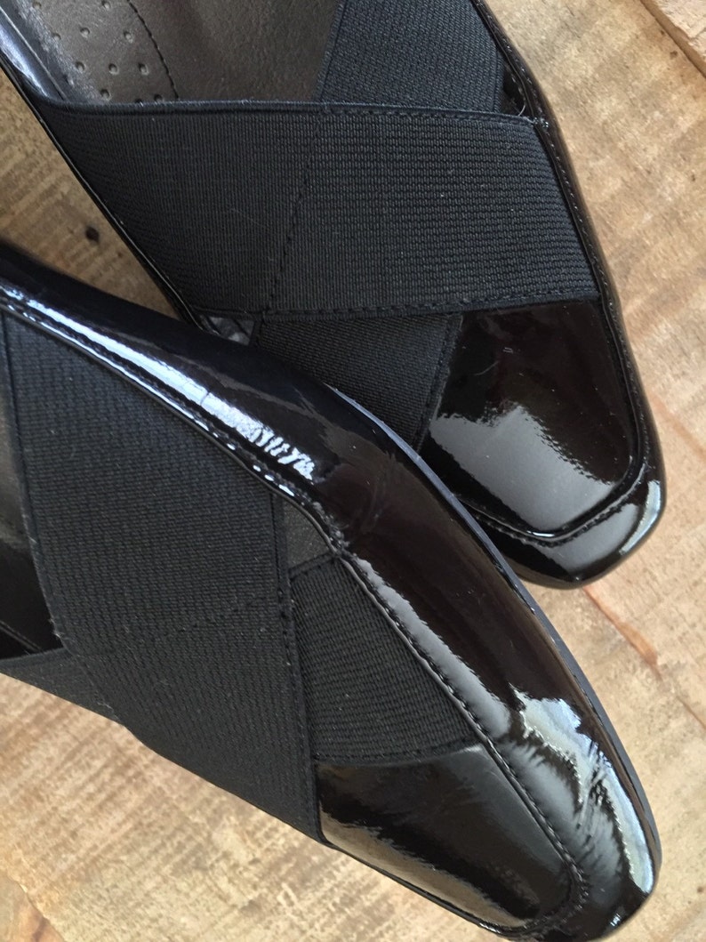 May include: A pair of black patent leather shoes with a criss-cross strap design. The straps are made of a black fabric.