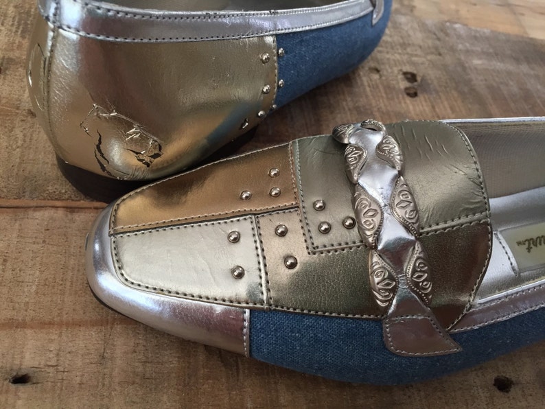 Puede incluir: Un par de zapatos de cuero plateado y dorado con detalles de denim. Los zapatos tienen un detalle de cadena plateada y tachuelas plateadas.