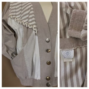 May include: A beige, striped, button-up jacket with a white and beige striped panel on the left side. The jacket has a V-neck and a label that reads "INFO" on the inside.