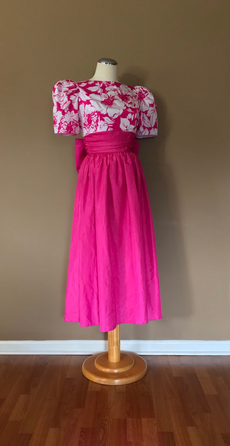 Puede incluir: Un vestido rosa con un corpi&ntilde;o de estampado floral blanco y una falda completa. El vestido tiene un lazo en la cintura y mangas cortas abullonadas.