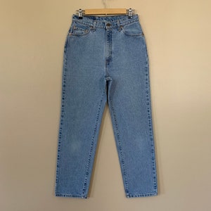 May include: Light blue denim jeans hanging on a wooden hanger. The jeans have a high waist, a button and zipper closure, and classic five-pocket styling. The denim appears to be a medium weight.