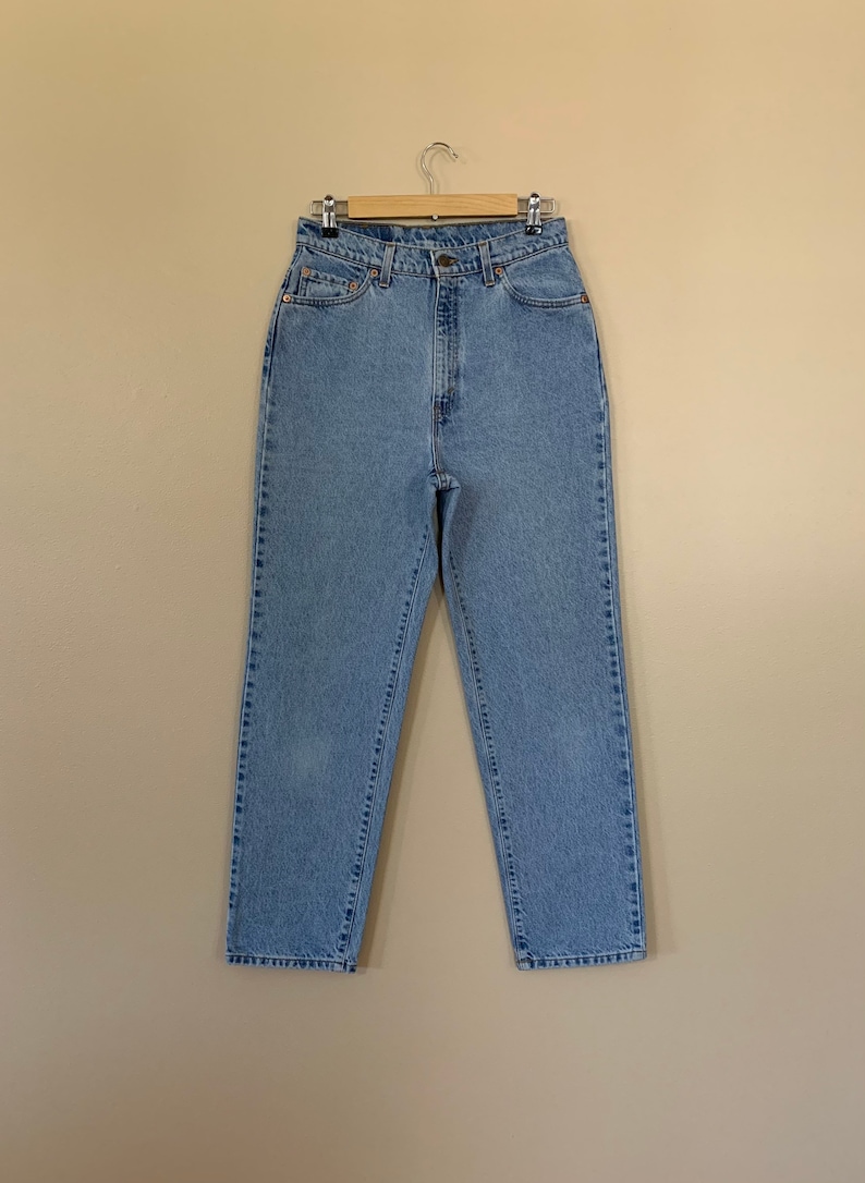 May include: Light blue denim jeans hanging on a wooden hanger. The jeans have a high waist, a button and zipper closure, and classic five-pocket styling. The denim appears to be a medium weight.