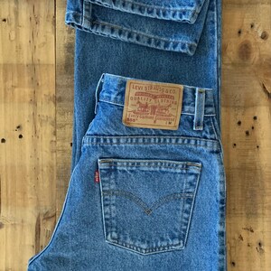 May include: Light blue denim jeans with a classic five-pocket design. The jeans feature a brown leather patch with the Levi Strauss & Co. logo and text. The jeans are folded on a wooden surface.