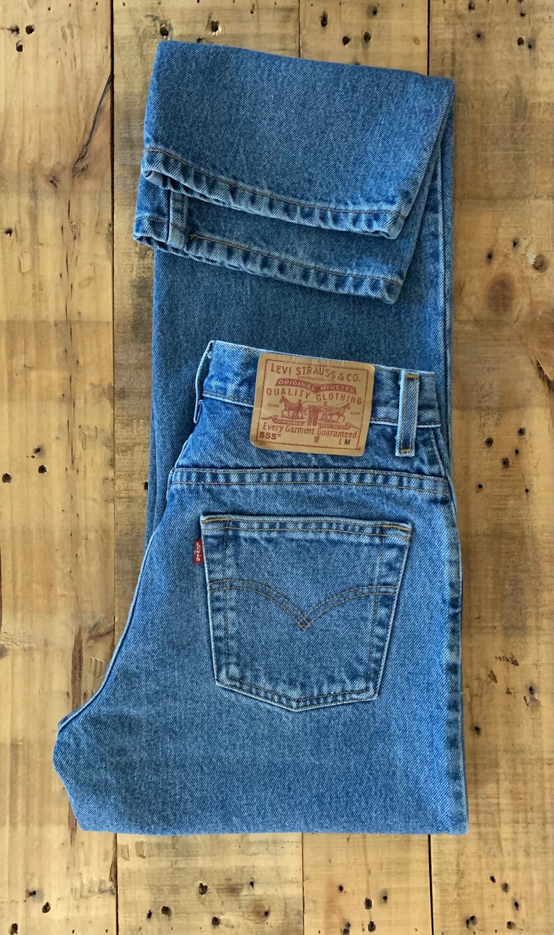 May include: Light blue denim jeans with a classic five-pocket design. The jeans feature a brown leather patch with the Levi Strauss & Co. logo and text. The jeans are folded on a wooden surface.