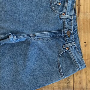 May include: A pair of light blue denim jeans with a button fly and a back pocket. The jeans are folded in half and laid on a wooden surface.