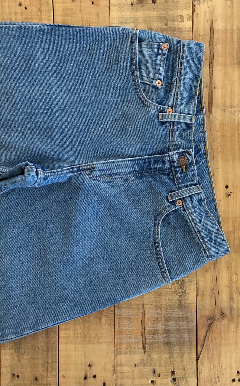 May include: A pair of light blue denim jeans with a button fly and a back pocket. The jeans are folded in half and laid on a wooden surface.
