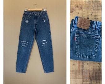 Könnte beinhalten: Eine blaue Jeans aus Denim mit abgenutzten Details. Die Jeans hat mehrere Risse und Löcher an den Knien und Oberschenkeln. Die Jeans hängt an einem Kleiderbügel vor einem beigefarbenen Hintergrund. Die Gesäßtasche der Jeans hat einen braunen Leder-Patch mit dem Text "Levi's" in Weiß.
