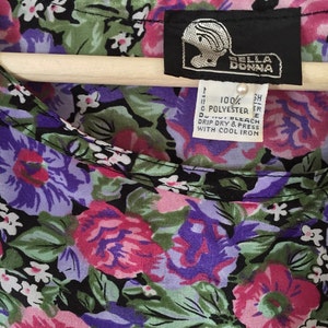 May include: A black and white floral print blouse with a pink, purple, and green color scheme. The blouse has a scoop neck and a label that reads "BELLA DONNA" and "100% POLYESTER".