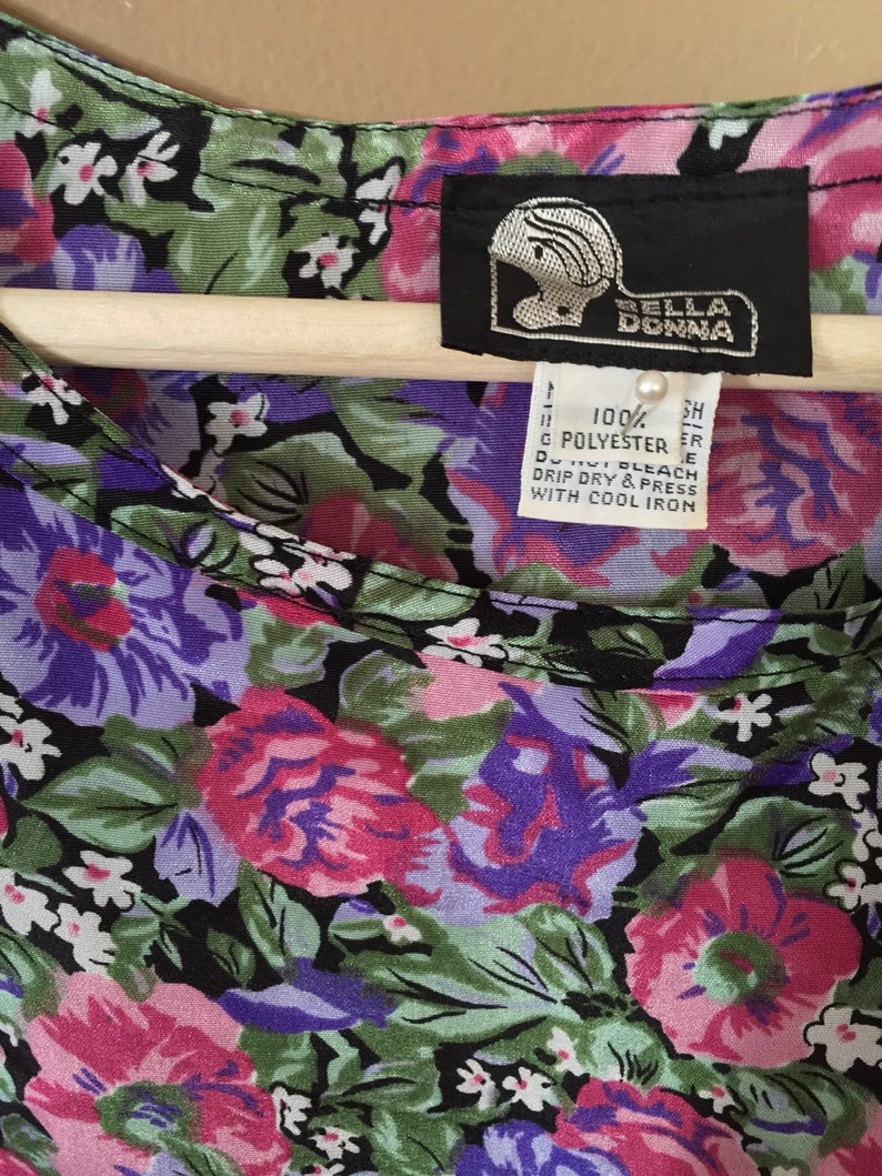 May include: A black and white floral print blouse with a pink, purple, and green color scheme. The blouse has a scoop neck and a label that reads "BELLA DONNA" and "100% POLYESTER".