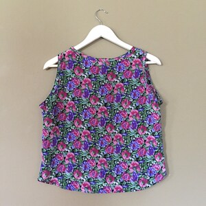 May include: A sleeveless floral print top with a pink, purple, and green color scheme. The top is hanging on a white hanger against a beige background.