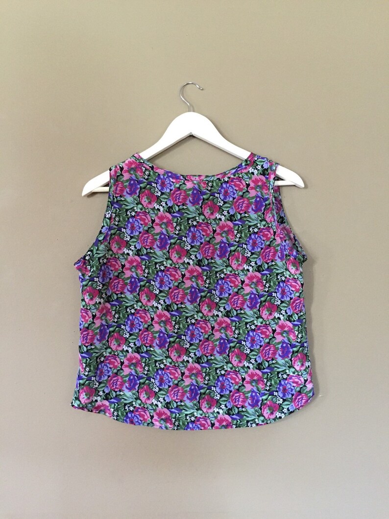 May include: A sleeveless floral print top with a pink, purple, and green color scheme. The top is hanging on a white hanger against a beige background.
