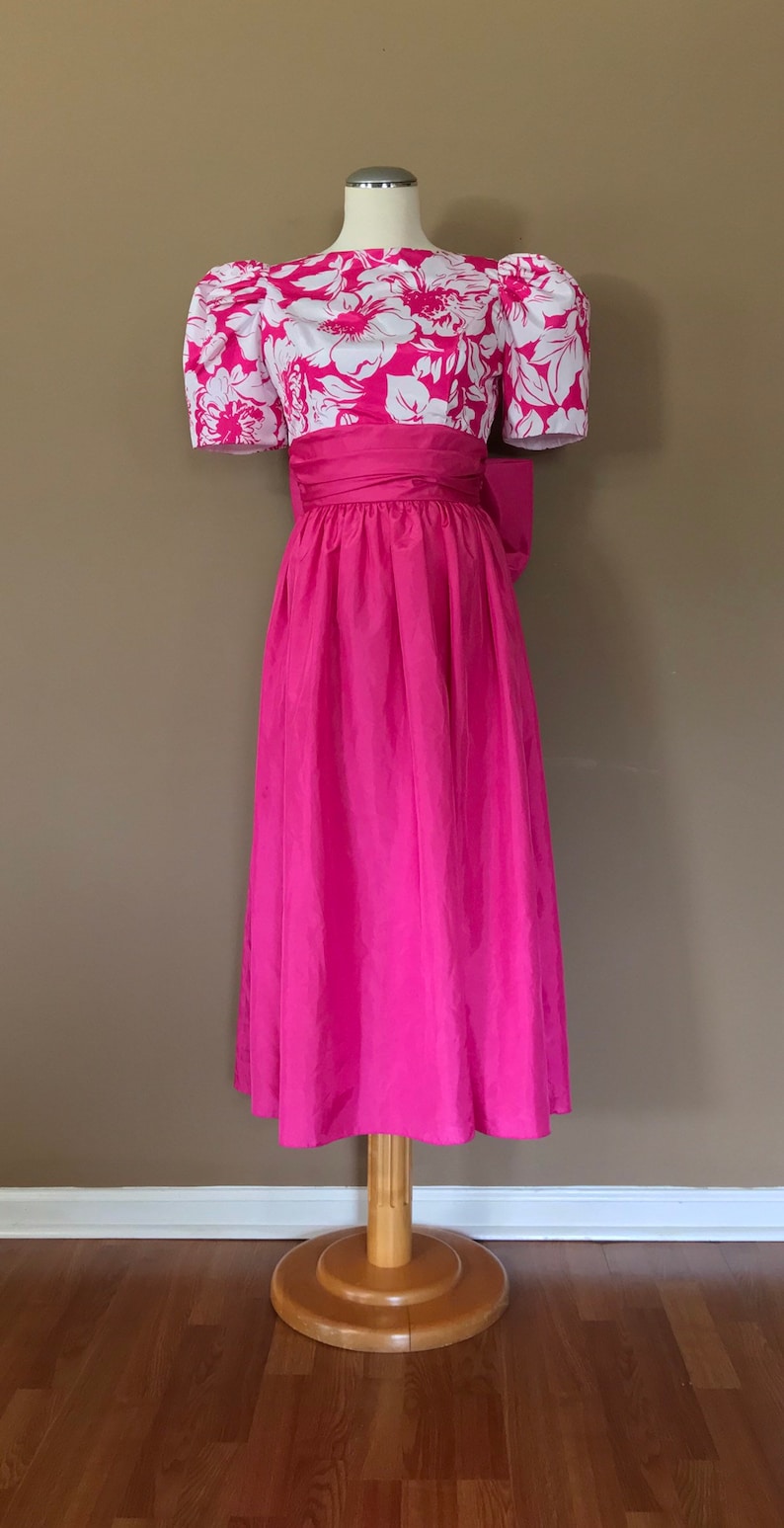 Puede incluir: Un vestido rosa con un corpi&ntilde;o de estampado floral blanco y una falda rosa lisa. El vestido tiene mangas abullonadas y un lazo en la cintura.