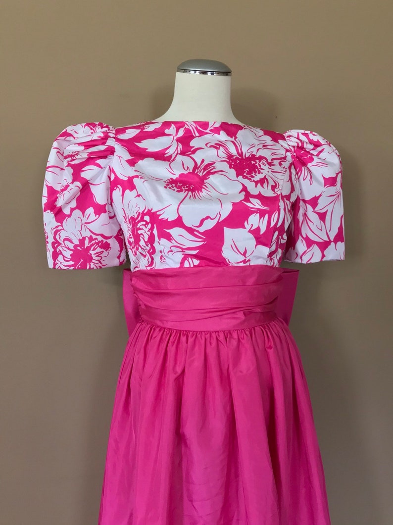 Puede incluir: Un vestido de estampado floral rosa y blanco con manga abullonada y una cinta a juego. El vestido tiene una falda completa y est&aacute; hecho de una tela ligera.