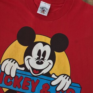 May include: Red t-shirt with a vintage Mickey Mouse graphic. Mickey is holding a blue and yellow sign that says "Mickey & Co."