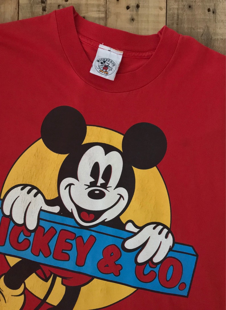 May include: Red t-shirt with a vintage Mickey Mouse graphic. Mickey is holding a blue and yellow sign that says "Mickey & Co."
