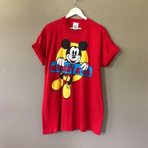 May include: Red short-sleeved t-shirt featuring a cartoon image of Mickey Mouse peeking over a blue and yellow sign that reads "Mickey & Co."