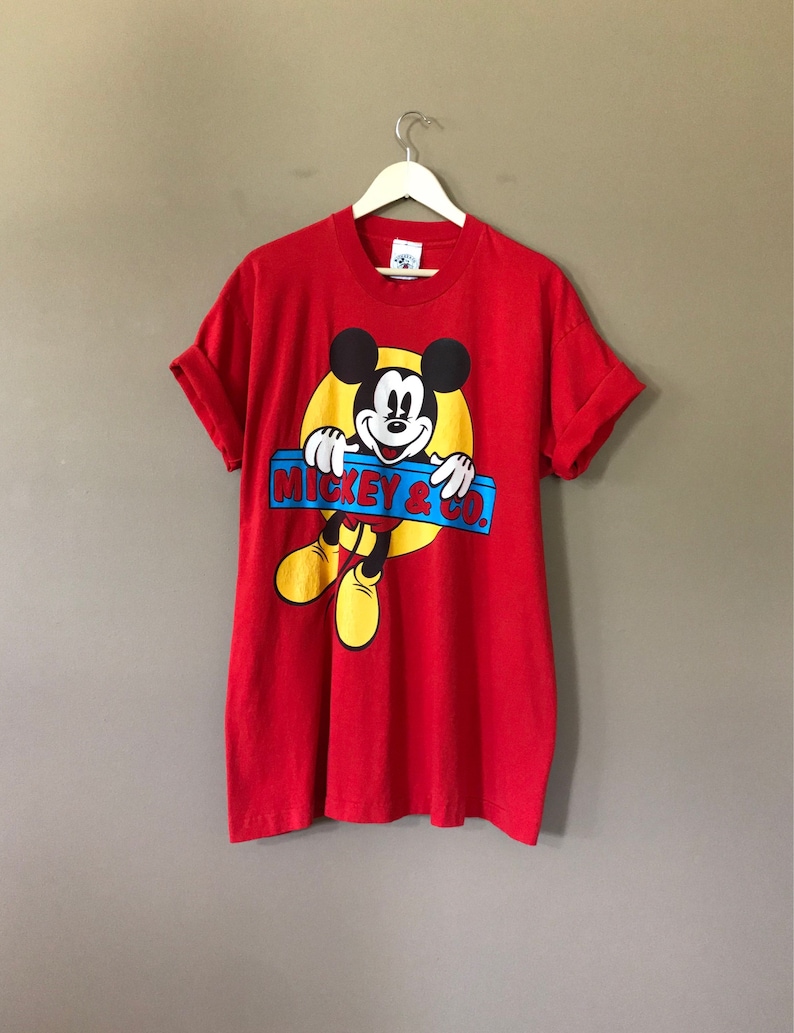 May include: Red short-sleeved t-shirt featuring a cartoon image of Mickey Mouse peeking over a blue and yellow sign that reads "Mickey & Co."