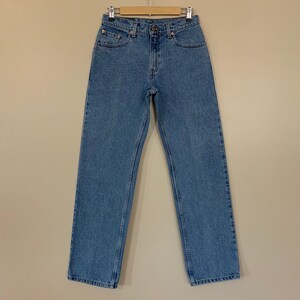 May include: A pair of light blue denim jeans hanging on a wooden hanger against a beige background.
