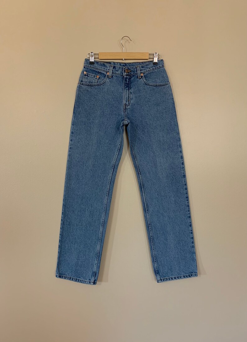 May include: A pair of light blue denim jeans hanging on a wooden hanger against a beige background.