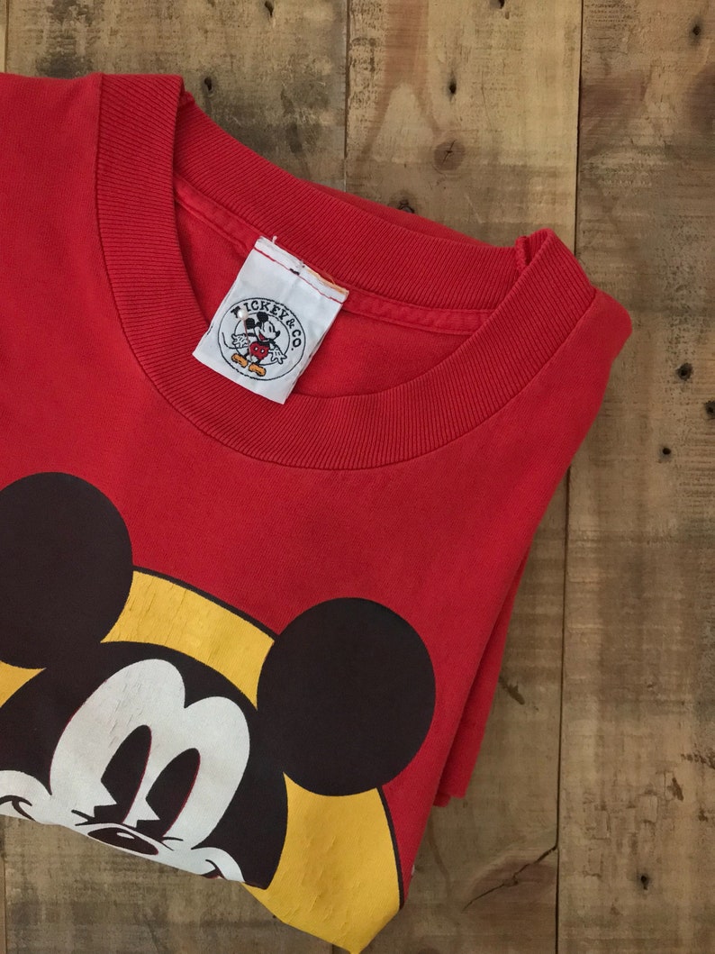 May include: Red t-shirt with a large Mickey Mouse graphic on the front. The shirt has a crew neck and a tag that says "Mickey & Co."