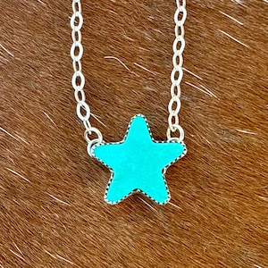 May include: A silver chain necklace featuring a turquoise star pendant. The star has a textured surface and a silver border. The necklace is displayed on a brown background.