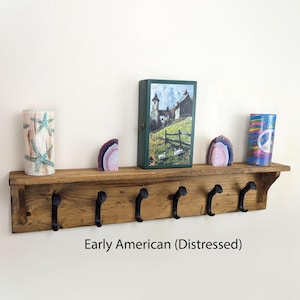May include: A distressed, Early American style wooden shelf with six black metal hooks. The shelf holds decorative items, including a cylindrical vase with starfish, a green box with a landscape painting, and two decorative stones.
