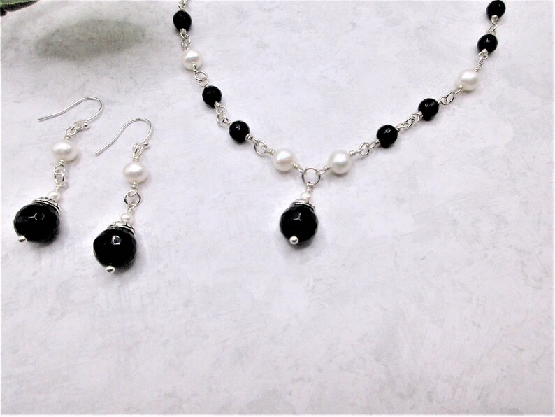 May include: A black and white pearl necklace with a black onyx pendant and matching black onyx earrings. The necklace and earrings are made with silver chain and beads.