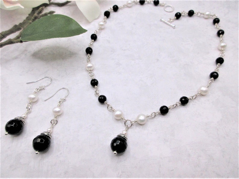 May include: A black and white beaded necklace with a black gemstone pendant and matching earrings. The necklace and earrings are made with silver chain and black and white beads.