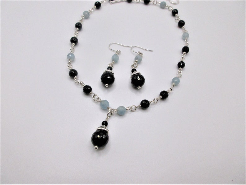 May include: A silver necklace and earrings set with black and light blue beads. The necklace has a pendant with a larger black bead.