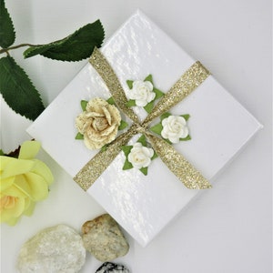 May include: A white gift box with a gold glitter ribbon and white and cream paper flowers. The box is sitting on a white surface with small rocks.