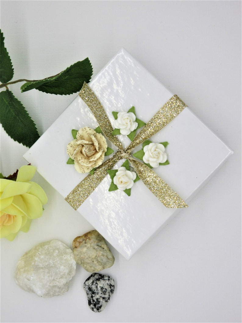May include: A white gift box with a gold glitter ribbon and white and cream paper flowers. The box is sitting on a white surface with small rocks.