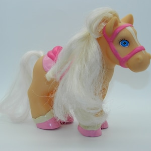 May include: A tan toy pony with a white mane and tail, and blue eyes. The pony has a pink saddle, bridle, and hooves. The toy is a vintage style, likely from the 1980s or 1990s.