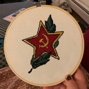 May include: Cross-stitch embroidery in a wooden hoop features a red star with a gold hammer and sickle emblem. The star is outlined in gold and surrounded by green leaves. The background is a cream-colored fabric.