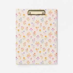Può includere: Una clipboard con un motivo floreale con piccoli fiori rosa, arancioni, gialli e blu su uno sfondo crema. La clipboard ha una clip in metallo dorato.