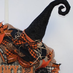 May include: A black witch's hat with a spiderweb pattern and a curled brim. The hat is decorated with a black and orange plaid bow and a black spider. The hat is part of a Halloween wreath.