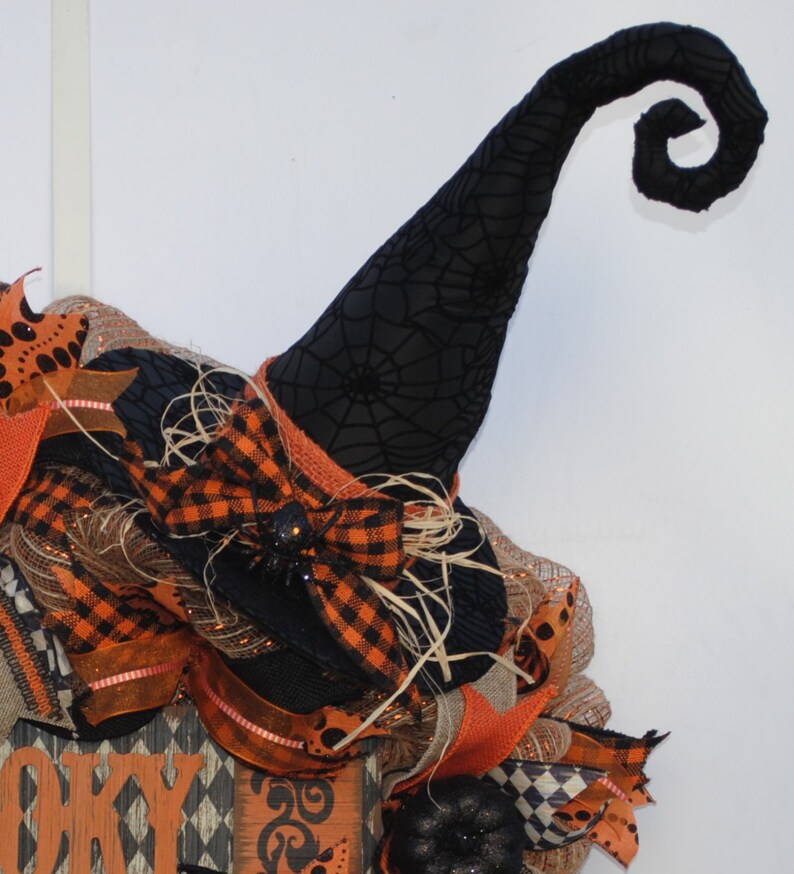 May include: A black witch's hat with a spiderweb pattern and a curled brim. The hat is decorated with a black and orange plaid bow and a black spider. The hat is part of a Halloween wreath.